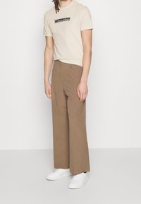 Osoba v béžovém tričku Calvin Klein, hnědých kalhotách s širokými nohavicemi a bílých teniskách, stojí proti jednoduchému světlému pozadí.