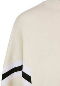 Světle béžová mikina s ribbovaným límcem, zdobená černobílými pruhy na rukávech. Měkká textura s volnějším střihem.