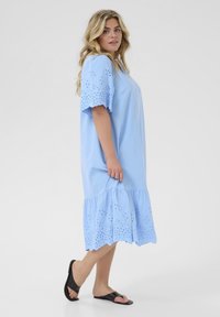 Lichtblauwe katoenen jurk met een uitlopende zoom, oogjesdetails bij de mouwen en onderkant, comfortabele pasvorm, gecombineerd met zwarte slidesandalen.