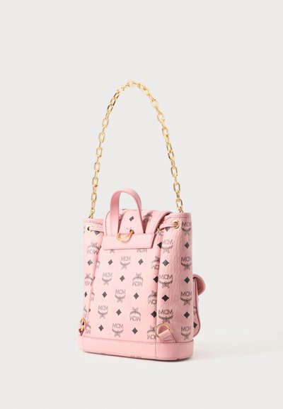 Sac à dos rose MCM avec imprimé monogramme noir, poignée chaîne dorée et bretelles en cuir sur un fond clair uni.