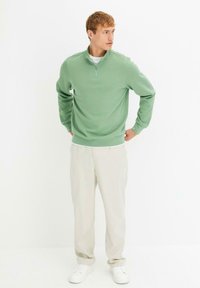 Maglione verde chiaro con collo zip realizzato in tessuto morbido, abbinato a pantaloni bianco sporco e sneakers bianche. Vestibilità comoda e texture liscia.