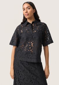 Svart spetsblus med krage, korta ärmar och blommönster. Tyget har en texturerad, genomskinlig look.