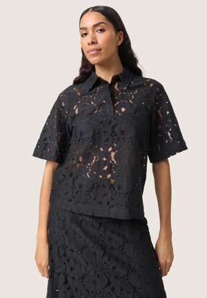 Blouse en dentelle noire avec un col, des manches courtes et des motifs floraux. Le tissu présente une apparence texturée et transparente.