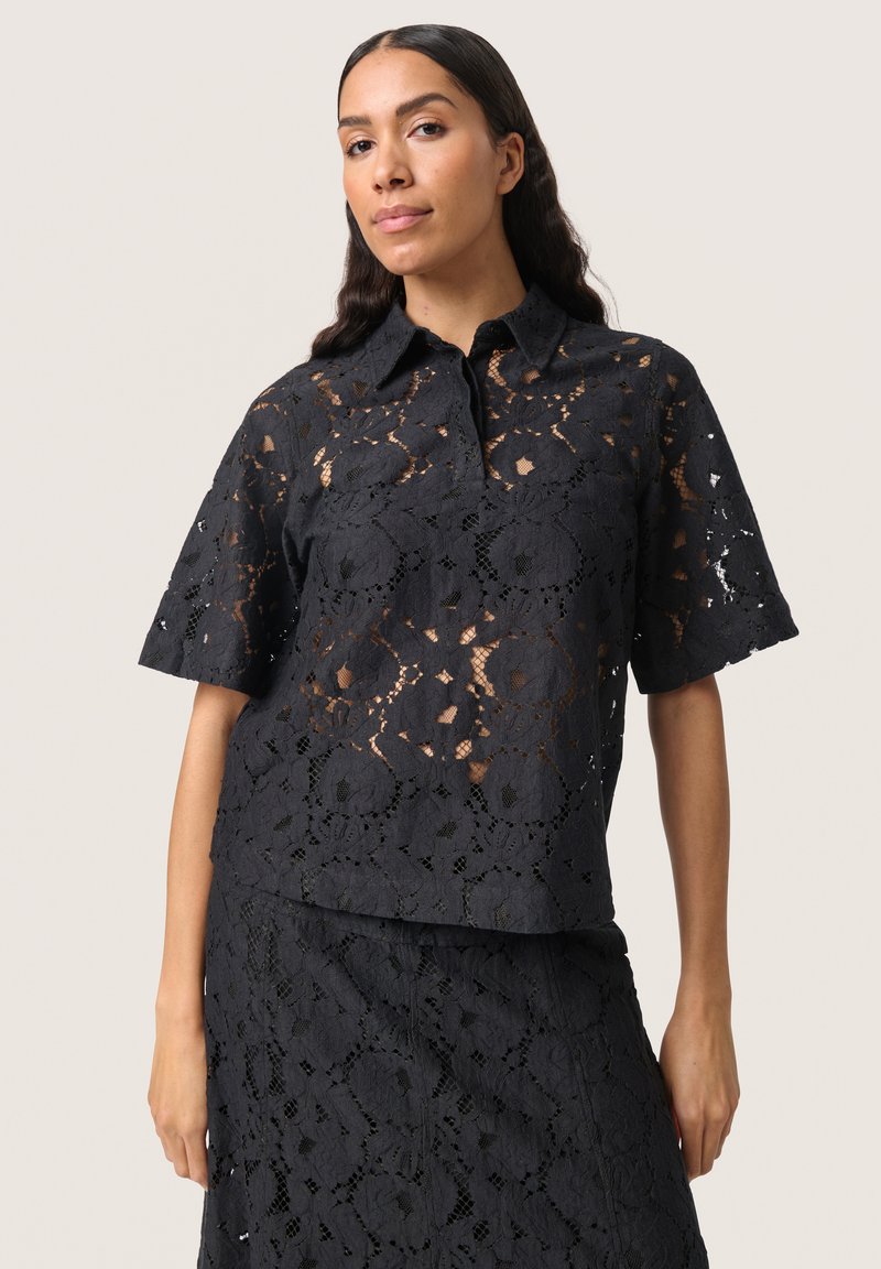 Svart spetsblus med krage, korta ärmar och blommönster. Tyget har en texturerad, genomskinlig look.