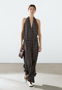Halter-neck jumpsuit in navy and brown checks, featuring an elastic waist and flowy wide legs; paired with a woven brown handbag and white shoes.