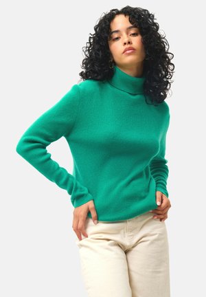 Pull en col roulé vert en tissu côtelé. Doté de manches longues et d'une coupe décontractée, associé à un pantalon de couleur claire.