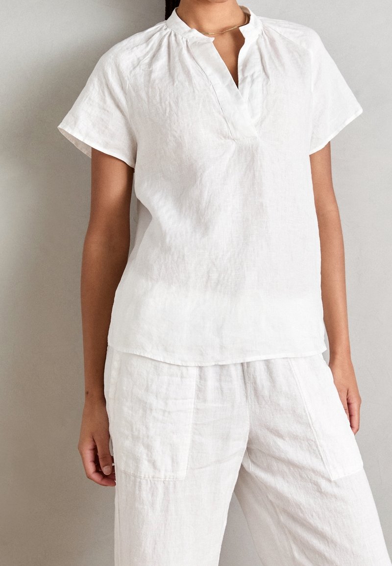 Haut en lin blanc avec une encolure en trou de serrure, des manches courtes et des détails froncés aux épaules. Associé à un pantalon en lin assorti.