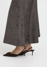 Talons hauts en suede marron à bride arrière avec un bout pointu, caractérisés par un détail en nœud et un talon fin. Portés avec une longue jupe gris texturé.