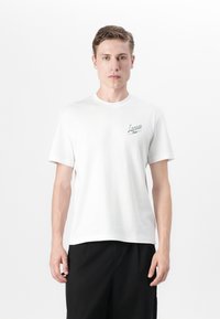 Balta medvilninė marškinėliai su mažuoju žaliu "Lacoste" logotipu kairiajame krūtinės šone, trumpomis rankovėmis ir apvaliu iškirpimu.