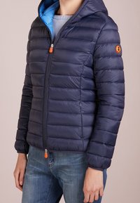 Tmavě modrá prošívaná péřová bunda s modrou podšívkou, předním zipem a oranžovým akcentem. Má kapuci a texturovanou, izolovanou látku.