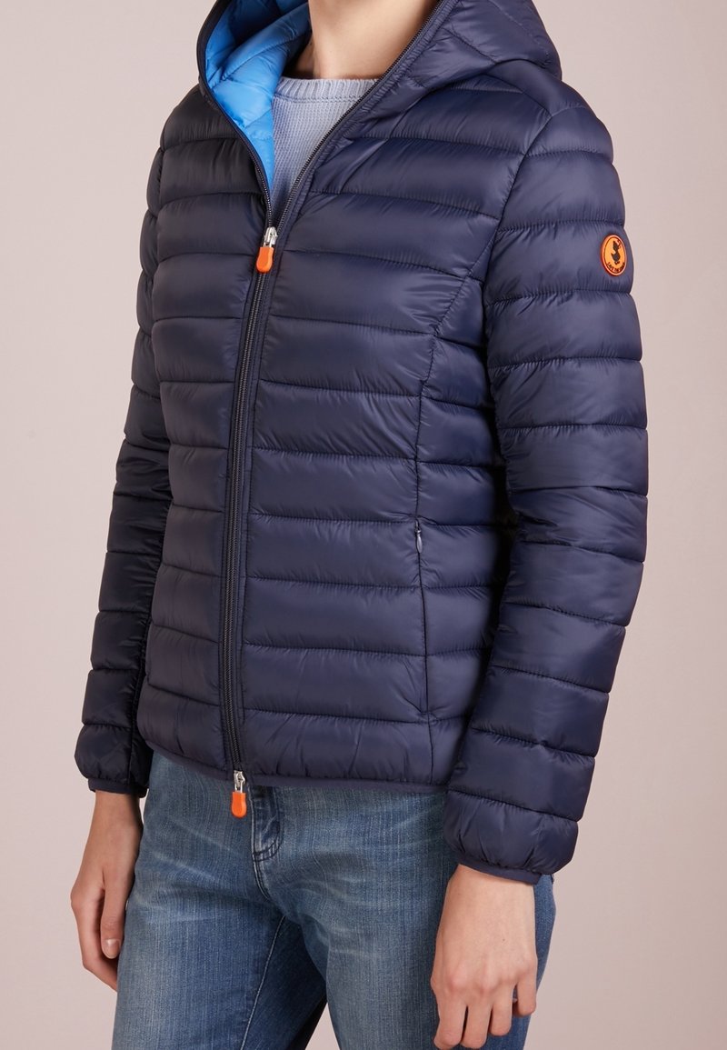 Tmavě modrá prošívaná péřová bunda s modrou podšívkou, předním zipem a oranžovým akcentem. Má kapuci a texturovanou, izolovanou látku.
