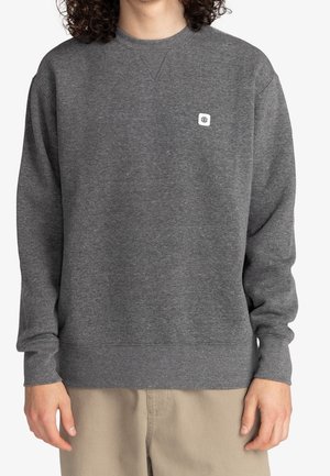 Osoba ubrana w ciemnoszary sweter typu crewneck z małym białym logo na lewej piersi oraz beżowe spodnie, stojąca na tle białej ściany.