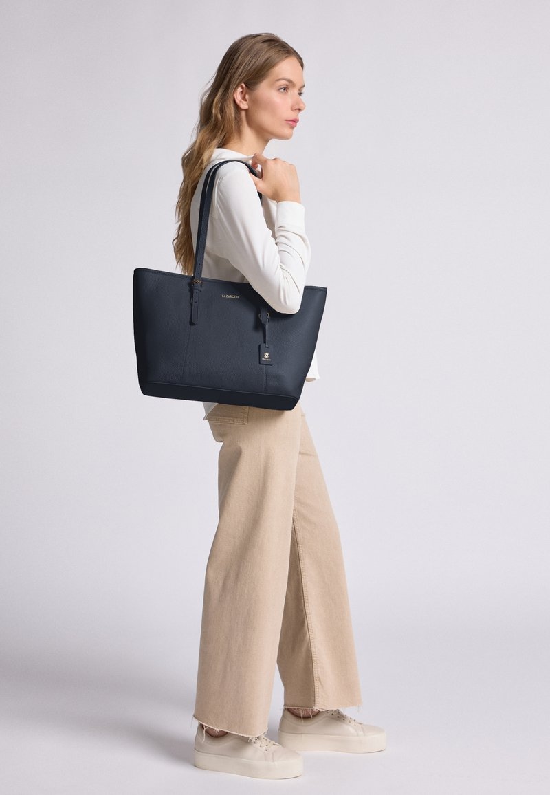 Borsa a spalla in pelle navy con manici lunghi, forma rettangolare, superficie testurizzata, dettaglio logo dorato e un piccolo tag attaccato per funzionalità.