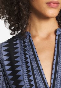 Blouse bleue à motifs avec des dessins géométriques noirs, un col en V, et une texture de tissu lisse. Le design comprend des motifs en zigzag et des triangles.