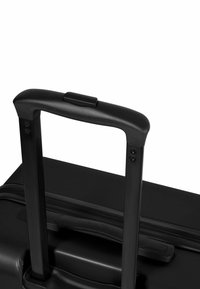 Valise noire à roulettes avec une surface texturée ; équipée d'une poignée rétractable, de vis en métal et d'une prise solide sur le dessus.