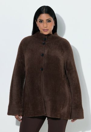 Kvinde med langt mørkt hår iført en brun fluffy knappet jakke og mørke bukser, stående mod en ensfarvet lys baggrund.