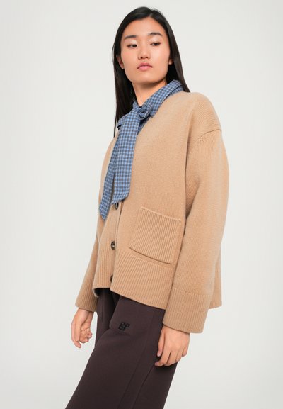 Bézs kötött kardigán, bordás mandzsettákkal és egy rakott zsebbel, kék kockás sállal a nyakban és sötétbarna nadrággal párosítva.