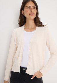 Lichtroze cardigan van zachte stof, met een v-hals, knopen aan de voorkant en geribbelde manchetten. Gelaagd over een wit shirt.