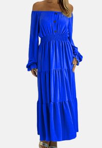 Kvinde iført en lang, off-shoulder kongeblå kjole med knapper og smocket talje, kombineret med sølvfarvede åbne sandaler.