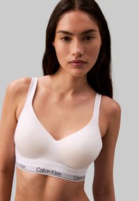 Witte katoenen bralette met v-halsontwerp, verstelbare bandjes en een zwarte logo-band aan de onderkant. Zachte textuur, minimalistische stijl.