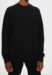 Schwarzer Pullover mit Rundhalsausschnitt und Raglanärmeln. Verfügt über ein strukturiertes, erhabenes Logodesign auf der Vorderseite für zusätzliche Details.