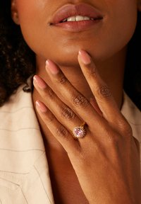 Gold ring with a large pink gemstone, surrounded by small clear stones. Hand with light pink nails rests near a neutral-colored fabric.