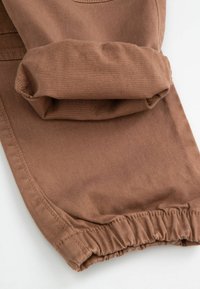 Brązowe bawełniane spodnie z podwiniętym mankietem, charakteryzujące się gładką fakturą i elastycznym ściągaczem na dole, podkreślające casualowe elementy designu.