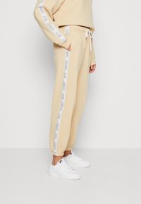 Beige joggers with white side stripe featuring printed text, elastic waistband with drawstring, and tapered cuffs. Worn with white sneakers.