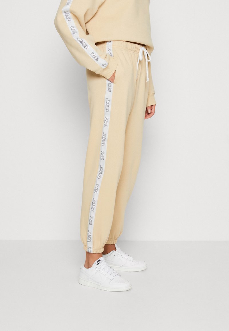 Beige joggers with white side stripe featuring printed text, elastic waistband with drawstring, and tapered cuffs. Worn with white sneakers.