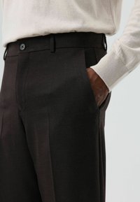 Donkere bruine op maat gemaakte broek met een gladde textuur, voorzien van voorzakken en sluiting met één knoop, gecombineerd met een lichte trui.