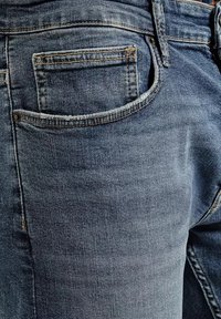 Ciemnoniebieskie dżinsy mają tylną kieszeń z wyszywaną krawędzią, widoczną teksturą i subtelnym wyblaknięciem. Wzmocnione szwy wzdłuż szwów.