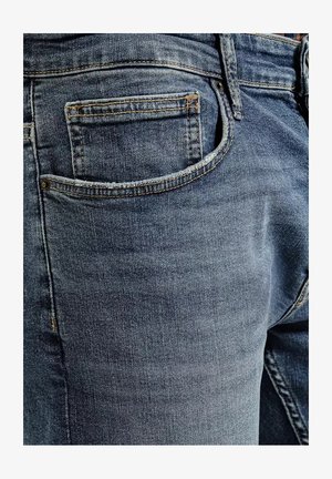 Ciemnoniebieskie dżinsy mają tylną kieszeń z wyszywaną krawędzią, widoczną teksturą i subtelnym wyblaknięciem. Wzmocnione szwy wzdłuż szwów.