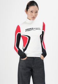 Duguljasti sportski top s bijelim tijelom, crvenim i crnim naglasima, te grafičkim printom. Uparen s tamnim trapericama.