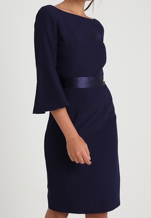 Femme portant une robe bleu marine à manches cloche et ceinture en satin assortie à la taille, sur un fond clair uni.