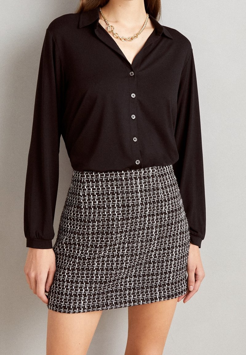 Femme portant une chemise noire à manches longues boutonnée, une mini-jupe à motifs noirs et blancs, et un collier chaîne en or.