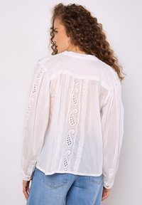 Weiße Bluse mit langen Ärmeln, Spitzenverzierungen und einer lockeren Passform. Sie verfügt über kunstvolle Stickereien und geknöpfte Manschetten. Aus leichtem Baumwollstoff.