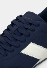 Σκούρα μπλε αθλητικά παπούτσια με υφασμάτινη επιφάνεια με υφή, διαθέτουν επίπεδα κορδόνια και λευκή διαγώνια ρίγα στο πλάι. Μινιμαλιστικός σχεδιασμός.