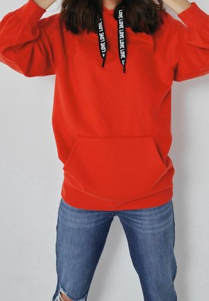 Personne portant un sweat à capuche rouge vif avec une poche avant et des cordons noirs imprimés "LOVE", associé à un jean bleu déchiré.