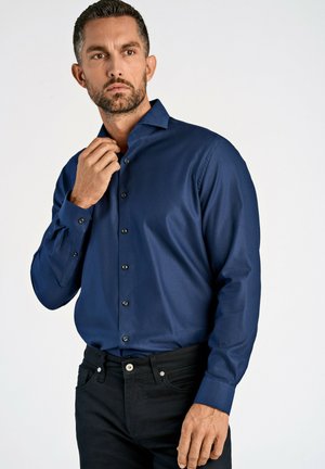 Navyblauwe langharige overhemd met een subtiele textuur, knoopsluiting en contrasterende zwarte knopen. Model draagt donkere jeans.