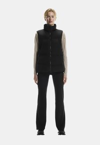 Sort puffer vest med høj krave og lynlås foran, sammen med en langærmet beige top og sorte bukser, der har en glat og slank tekstur.