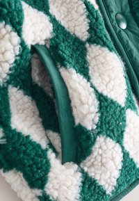 Tessuto a quadretti verdi e bianchi, con una superficie soffice, strutturata e pelosa. Fodera verde e dettaglio con bottone completano il design.