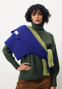 Groene coltrui gelaagd met een blauwe ribbeltrui die over de schouder is gedrapeerd, met een zachte textuur en een casual ontwerp.