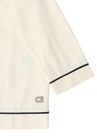 Jasno-beżowa koszula z długim rękawem z czarnym wykończeniem przy mankietach i dolnej krawędzi, z małym czarnym logo "CE Reflection" blisko dolnej krawędzi.