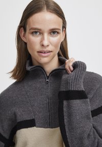 Pull en tricot gris avec un col zippé, featuring des blocs de couleur noir et beige audacieux, des poignets côtelés et un tissu doux et texturé.