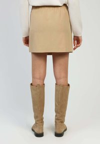 Jupe mini beige avec une taille élastique, associée à des bottes en daim beige montantes. Texture lisse, design simple et détails minimalistes.