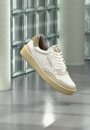 Zapatilla AllSaints blanca y beige flotando sobre un suelo reflectante con pared de bloques de vidrio y columna en el fondo.