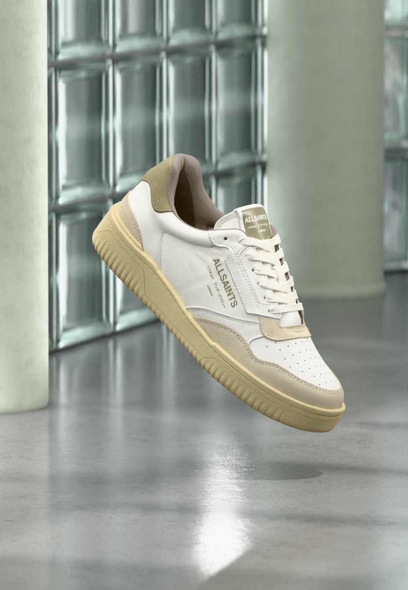 Λευκό και μπεζ sneaker AllSaints να αιωρείται πάνω από ανακλαστικό πάτωμα με γυάλινο τούβλο στον τοίχο και κολώνα στο φόντο.