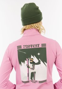 Rožinė ilgomis rankovėmis marškiniai su grafikos atspaudu, kuriame parašyta "PROTEST", ir slidinėjimo tema iliustracija ant nugaros. Žalia megzta kepurė.
