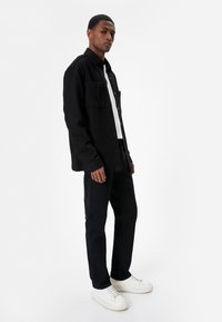 Chaqueta negra de botones sobre una camisa blanca, combinada con pantalones negros y zapatillas blancas. Presenta un corte relajado y detalles de bolsillo.