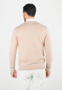 Pull beige clair à manches longues, col rond et poignets côtelés. Texture lisse, design ajusté, porté par-dessus une chemise blanche à col.